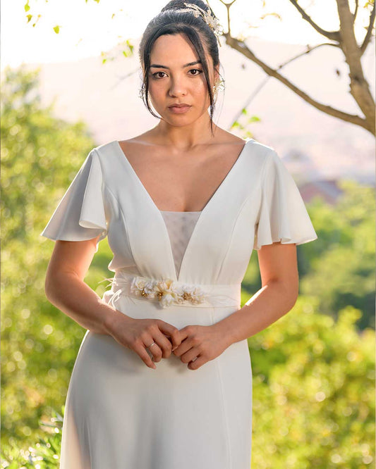 Vestido blanco largo para novia civil o invitadas de fiestas de blanco, con mangas, escote en V, cintuon con aplique floral. La modelo se encuentra en un parque.