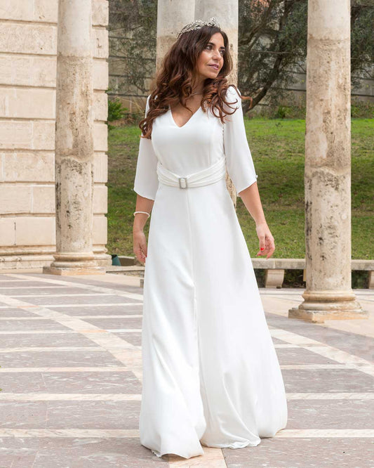 Vestido largo blanco de novia de mangas 3/4, cinturón con aplique cuadrado plateado y escote de pico.