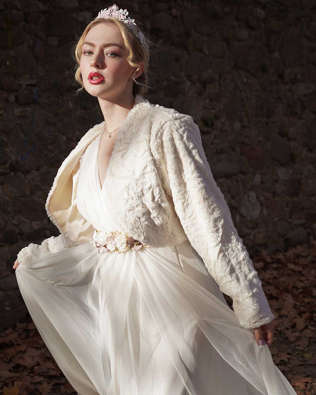 Chica vestida de novia con una torera de pelo blanca y un cinturón de flores, camina con una corona blanca mientras mira a la cámara.