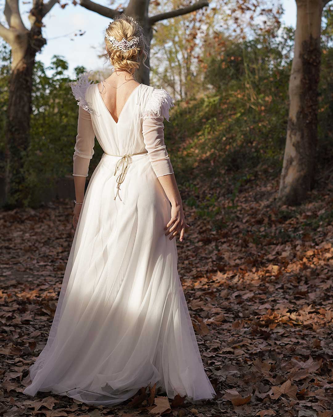 Chica rubia de espaldas lleva un vestido blanco de novia, el cual tiene plumas en los hombros, mangas de gasa 3/4 y un cinturón dorado.