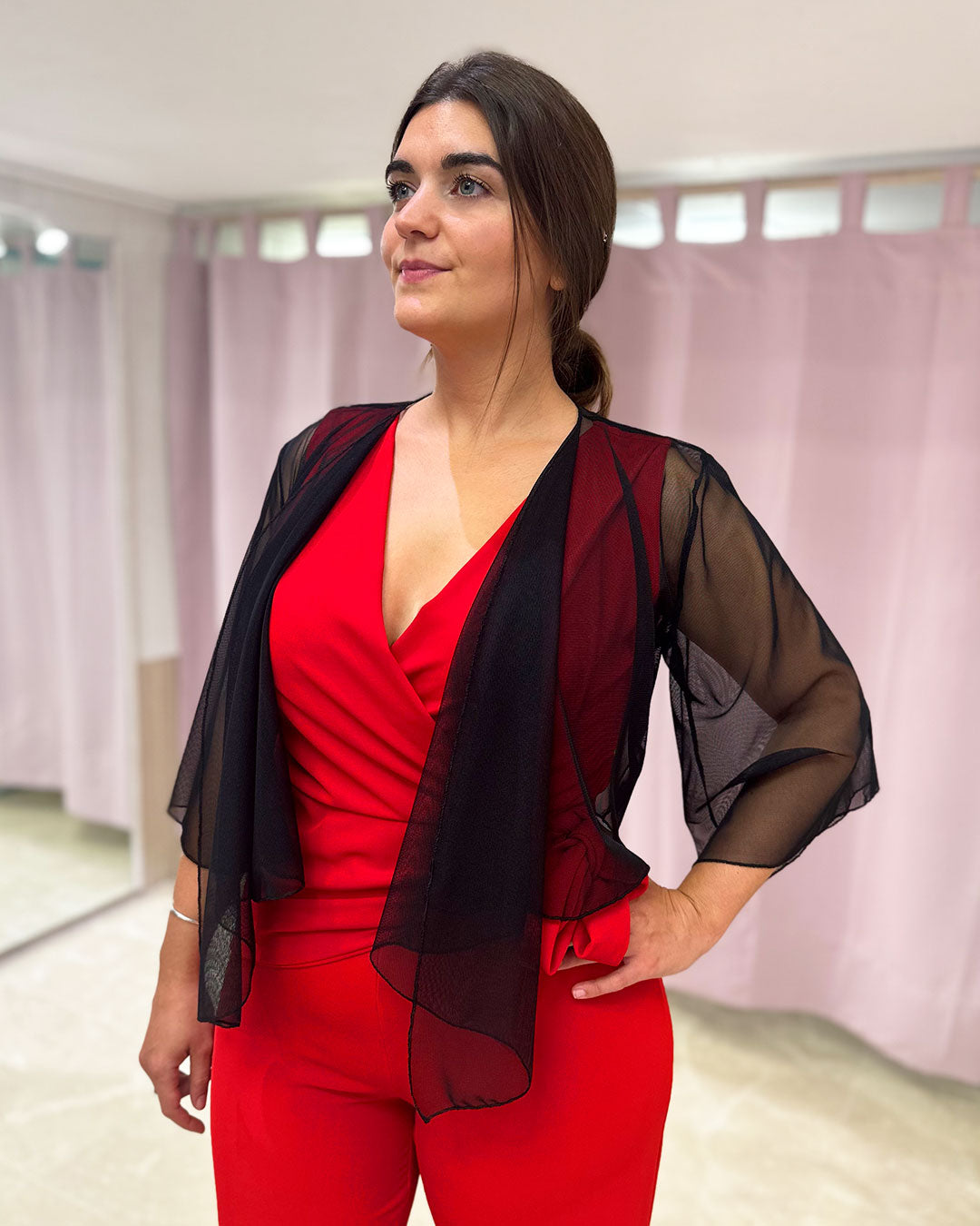 Chica dentro de un local lleva un mono rojo combinado con un bolero color negro que tapa sus brazos hasta la antemano. Su tela es de gasa transparente.